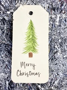 a tag with a christmas tree painted on it sitting in front of silver tinsel