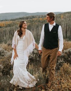 a bride and groom holding hands walking through tall grass with trees in the back ground