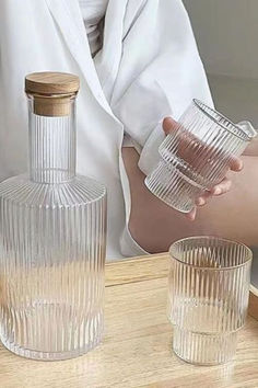 a woman holding two glasses next to an empty glass bottle on a wooden table with another glass in front of her