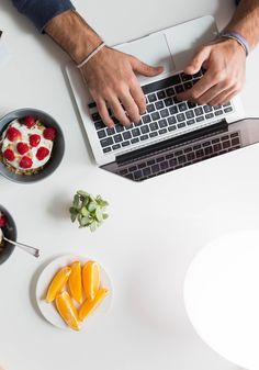 a person typing on a laptop computer surrounded by fruit and desserts in small bowls