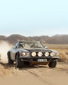 a black car driving down a dirt road in the middle of some grass and bushes