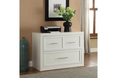 a white dresser with two drawers in front of a painting and vase on the wall