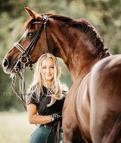 a beautiful blond woman standing next to a brown horse in a field with trees behind her