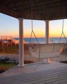 a white swing chair sitting on top of a wooden deck next to the ocean at sunset