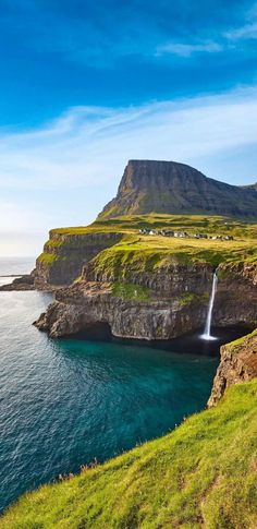 a waterfall in the middle of a body of water next to a green hill and blue sky