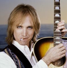 a man holding an acoustic guitar in his right hand and looking at the camera while standing next to the ocean