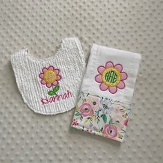 two bibs with flowers on them sitting next to each other, one has a monogrammed applique