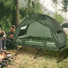 two men sitting next to each other near a campfire with a tent in the background