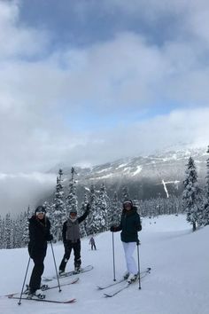 three people on skis standing in the snow with trees behind them and clouds above