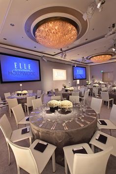 a large banquet room set up with white chairs and round tables in front of two big screens