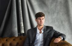 a man sitting on top of a brown couch in front of a gray wall with curtains