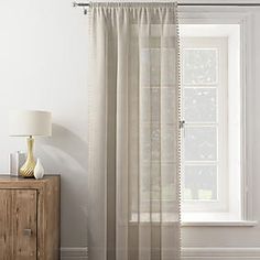 a white curtain hanging on the side of a window next to a dresser with a lamp