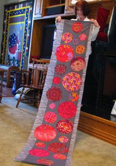 a woman holding up a large quilt in front of a fire place with many circles on it