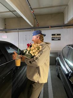 a young man holding flowers next to a car in a parking garage with a drink