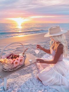 a woman in a white dress and hat sitting on the beach with a basket of fruit