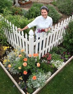 a woman standing next to a white picket fence with flowers growing in the garden below