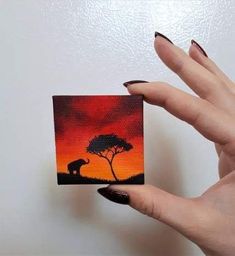 a woman's hand holding up a small painting with an elephant and tree in the background