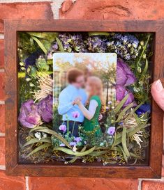 a person holding up a photo in front of a brick wall with purple flowers and greenery