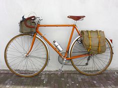 an orange bicycle parked next to a white wall with a bag on it's back