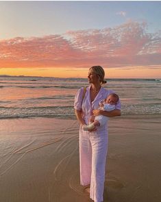 a woman holding a baby on the beach at sunset