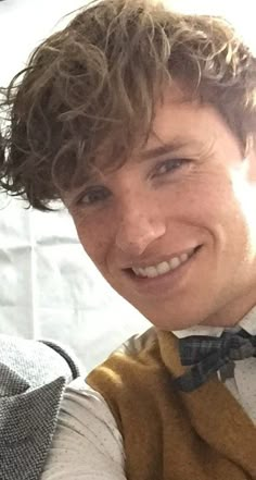 a young man with curly hair wearing a bow tie and sweater vest smiling at the camera