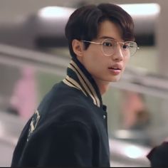 a young man wearing glasses and a black jacket is standing in an airport terminal looking at the camera