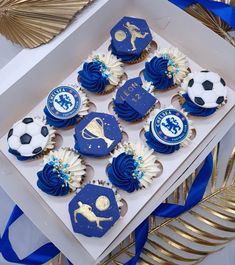 cupcakes decorated with blue icing and soccer balls are displayed in a box