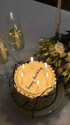 a birthday cake with lit candles on it sitting next to some flowers and wine glasses