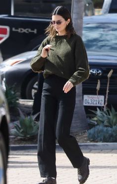 a woman in green sweater and black pants walking down the street with her cell phone