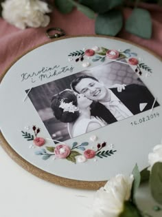 an embroidered photo frame with flowers and greenery around it, next to a bouquet of white roses