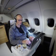 a man sitting in an airplane with headphones on working on his laptop and drinking coffee