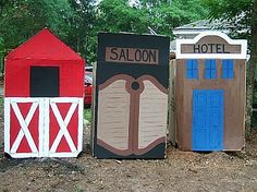 three small wooden outhouses painted in different colors and sizes, with trees behind them