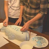 two people standing over a cake on top of a wooden table with white frosting