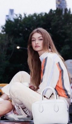 a woman sitting on top of a car holding a white purse and looking at the camera