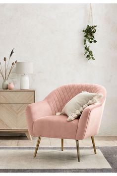 a pink chair with pillows on it in front of a white wall and wooden dresser