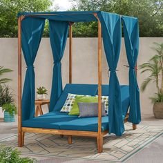 an outdoor daybed with blue curtains and pillows on it's sides, in front of a wall