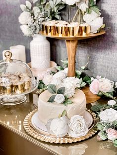 a wedding cake with white flowers and greenery sits on a gold platter next to two tiered cakes