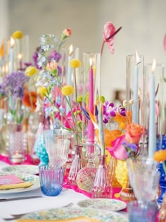 the table is set with many colorful vases and flowers in glass bottles, candles, and napkins