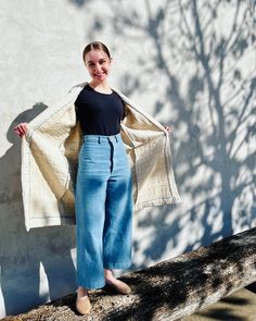 a woman standing next to a tree with her coat over her shoulders and arms outstretched