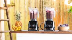 two blenders sitting on top of a wooden table next to a bowl of fruit