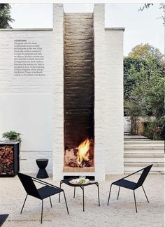 an outdoor fireplace with two chairs and a table next to it in front of a white wall