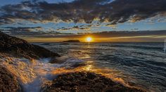 the sun is setting over the ocean with waves crashing on rocks in front of it