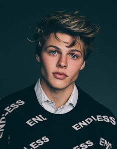 a young man with blue eyes wearing a black sweater and looking at the camera while standing in front of a dark background