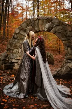 two women dressed in medieval clothing standing next to each other near a stone arch and trees