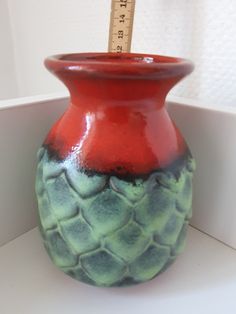 a red and green vase sitting on top of a white shelf next to a ruler