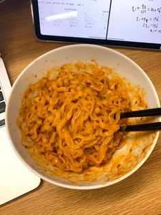 a bowl of noodles with chopsticks in front of a laptop