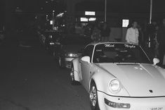 black and white photograph of cars parked on the side of the road with people standing around