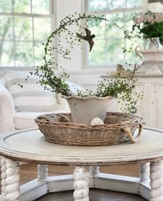 a white table topped with a basket filled with flowers and greenery next to a window