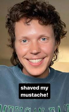 a young man with curly hair smiling and wearing a blue shirt that says shaved my mustache