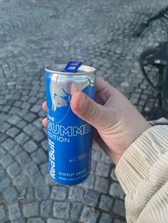 a person holding up a can of energy drink in their left hand on the cobblestone street